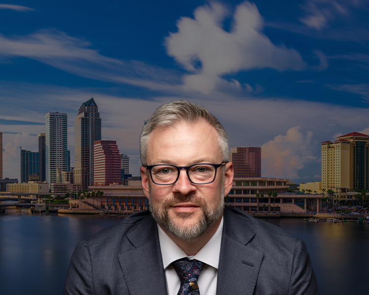 Professional portrait of Jason M. Mayberry, in a suit and tie, set against a panoramic background of Tampa's city skyline at dusk.