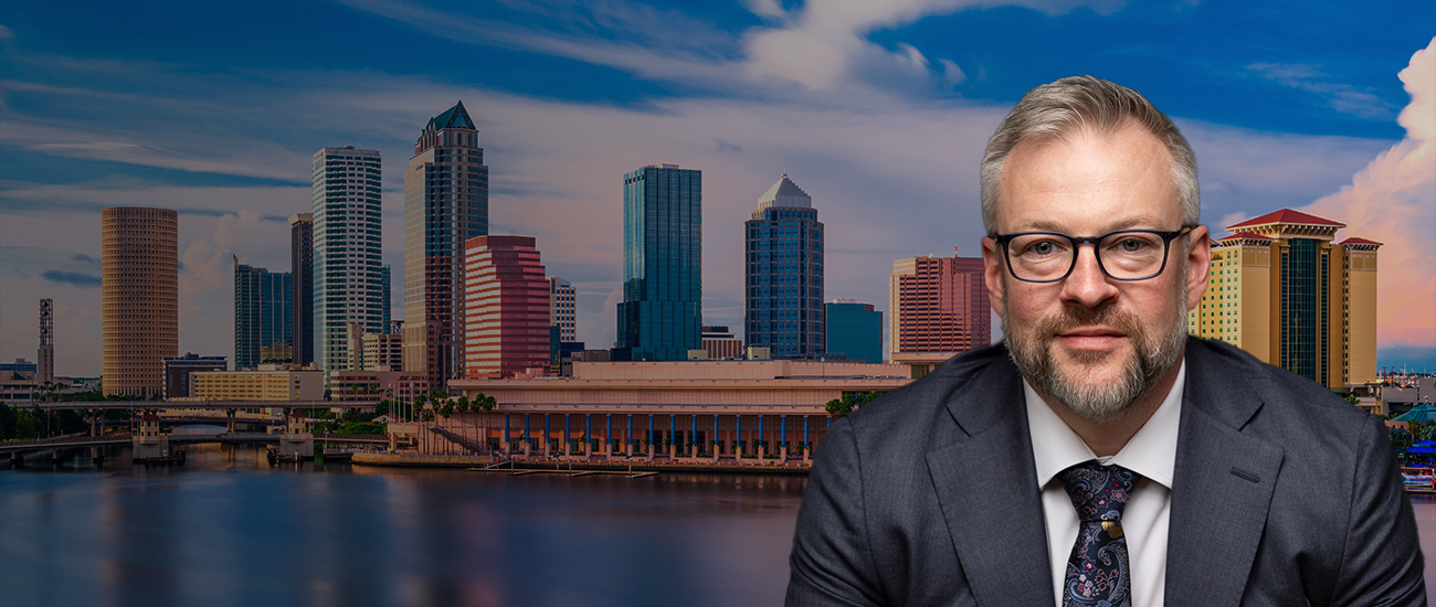 Professional portrait of Jason M. Mayberry, in a suit and tie, set against a panoramic background of Tampa's city skyline at dusk.
